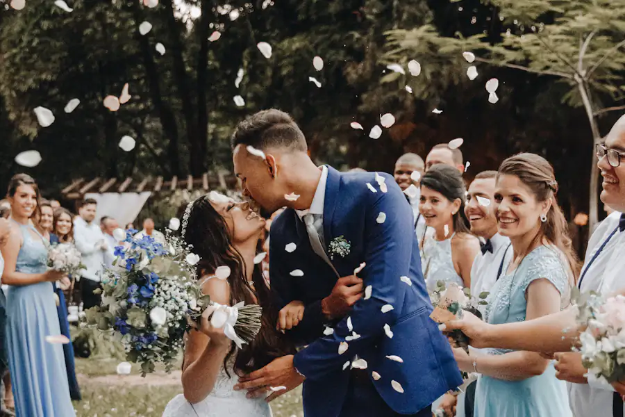 Noivos se beijando sob uma chuva de pétalas, cercados pelos convidados